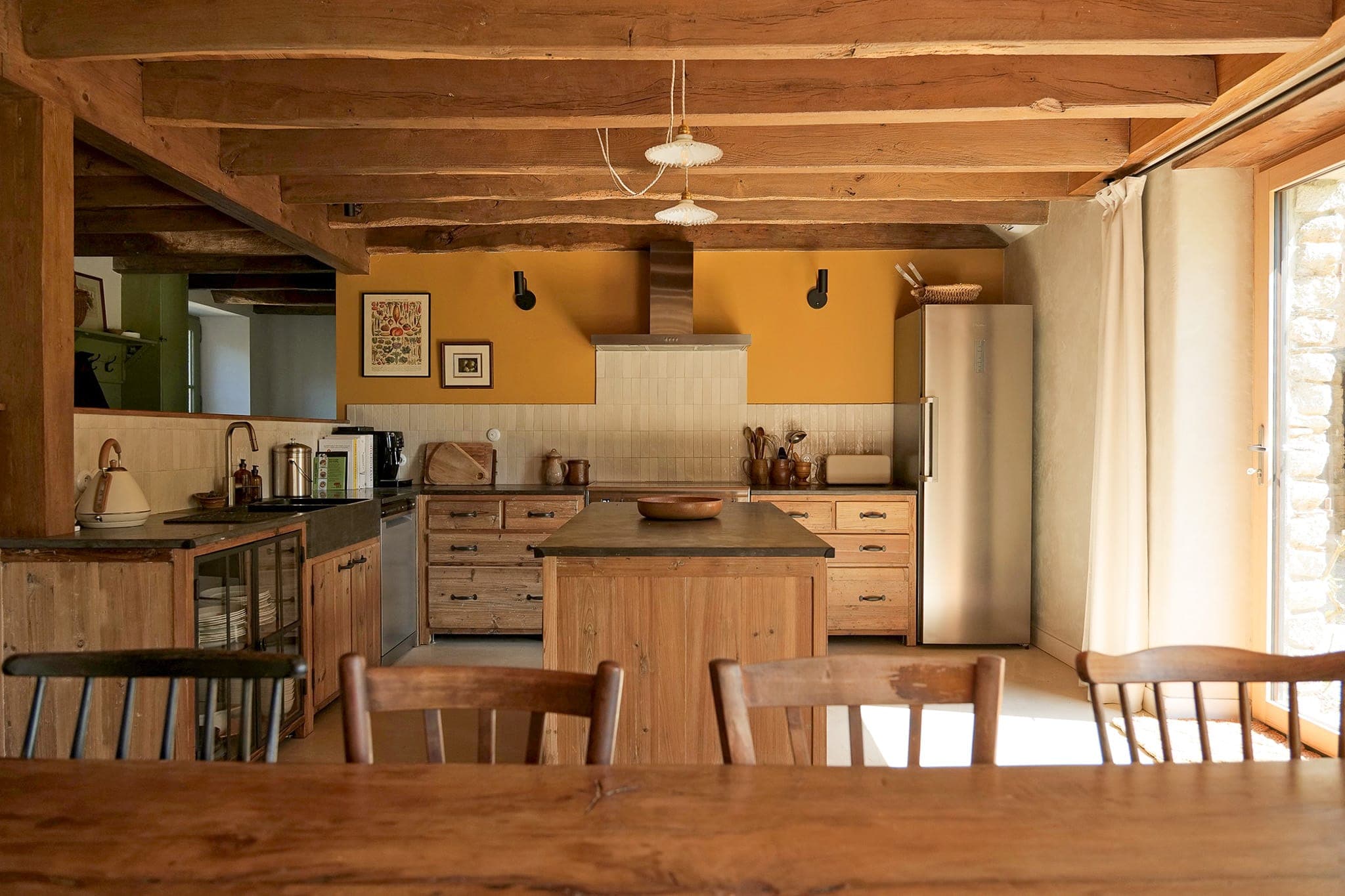 A rustic kitchen with exposed beams, wooden cupboards, central island and stainless steel refrigerator. Sunlight streams in through a window, illuminating this warm, cosy space. Four wooden chairs are arranged around a dining table in the foreground.