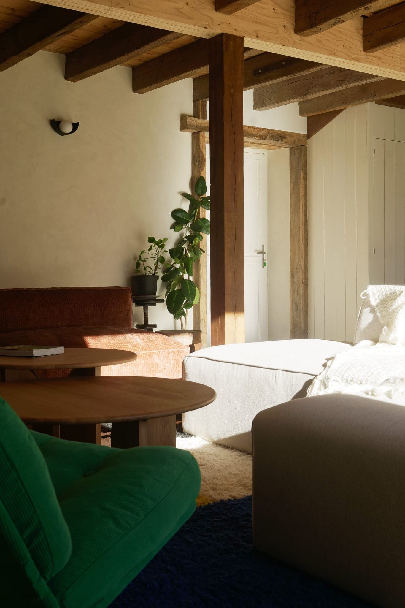 A comfortable, sunny room with wooden beams, a green armchair, a brown sofa, round wooden tables, a bed with white bedding, a potted plant and a white door in the background.