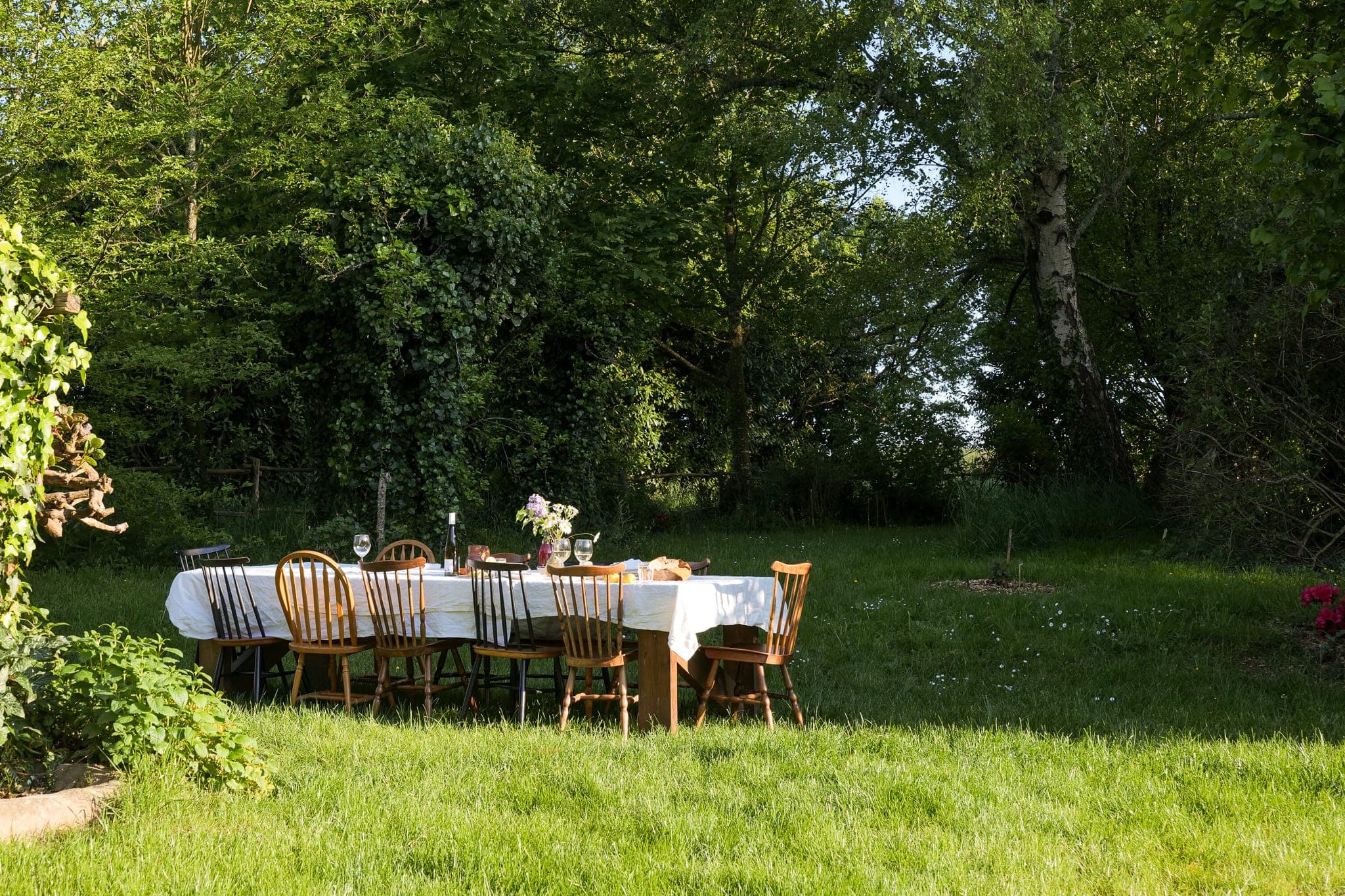 A long outdoor dining table with white tablecloth, wooden chairs and floral centerpiece sits on the grass in a lush, green garden surrounded by trees and sunshine.