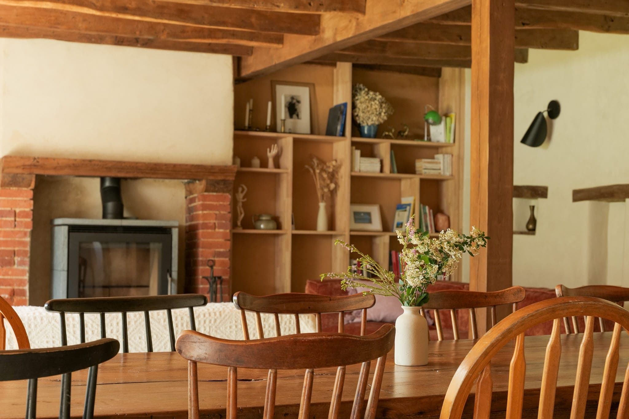 A warm dining room with a wooden table and mismatched chairs, a vase of flowers in the center, a wood-burning stove and built-in shelves with books and decorative objects in the background.