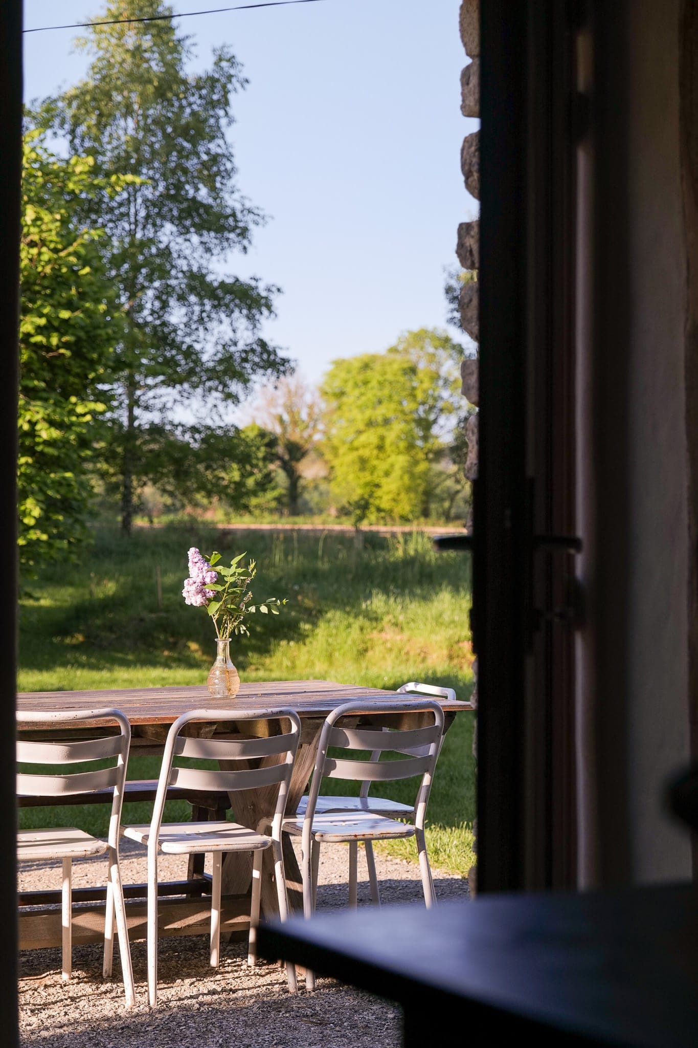 A wooden outdoor table and four metal chairs sit on a terrace, supporting a glass vase of purple flowers. Lush green trees and lawns loom under a clear blue sky in the background.