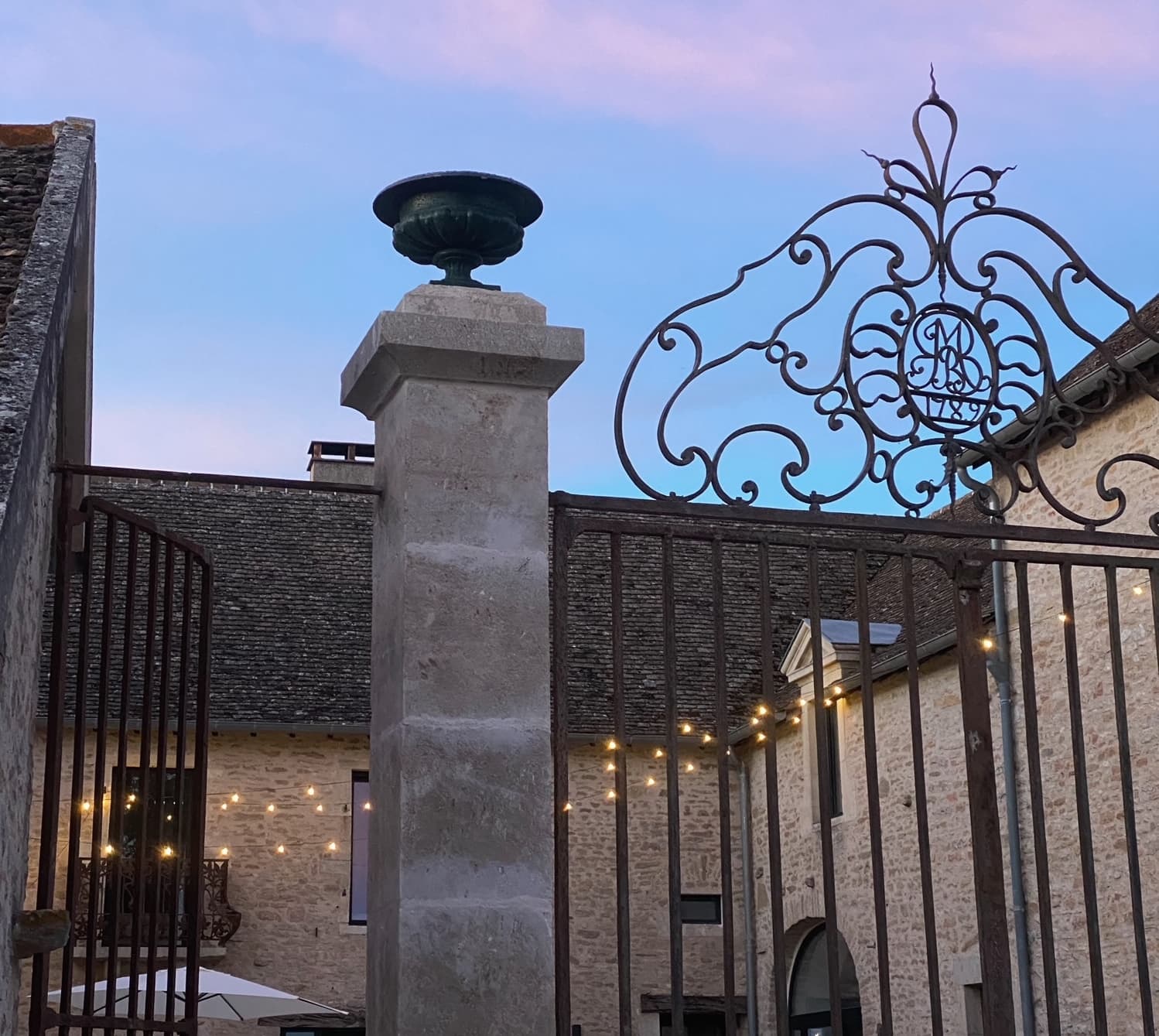 A monogrammed iron gate stands between stone buildings at sunset. Garlands of lights adorn the courtyard, and the sky is tinged with pink and blue.