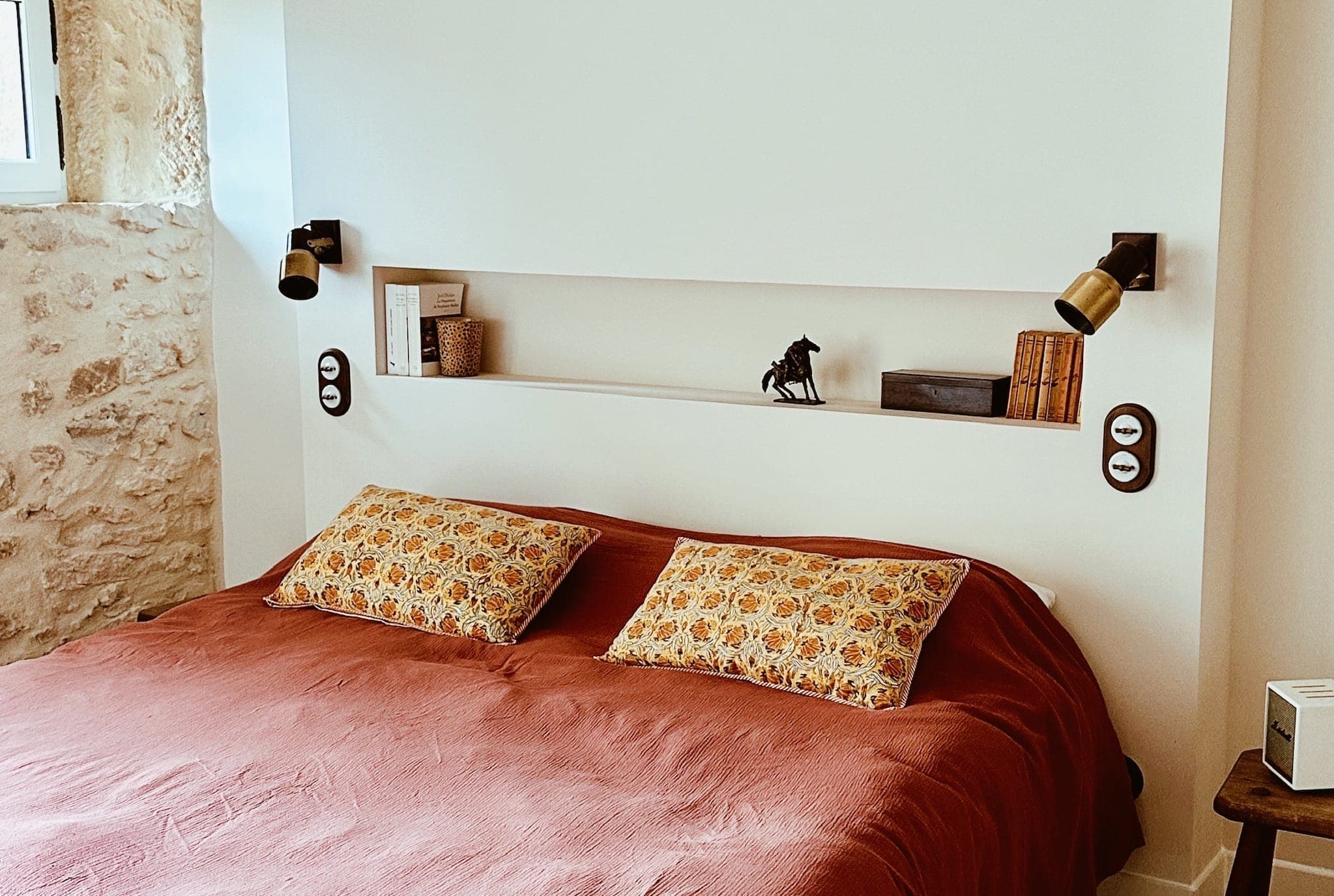 A cozy room with a double bed covered with a rust-colored blanket and two patterned pillows, built-in wall shelves with books and a horse figurine, wall lamps and a stone accent wall.