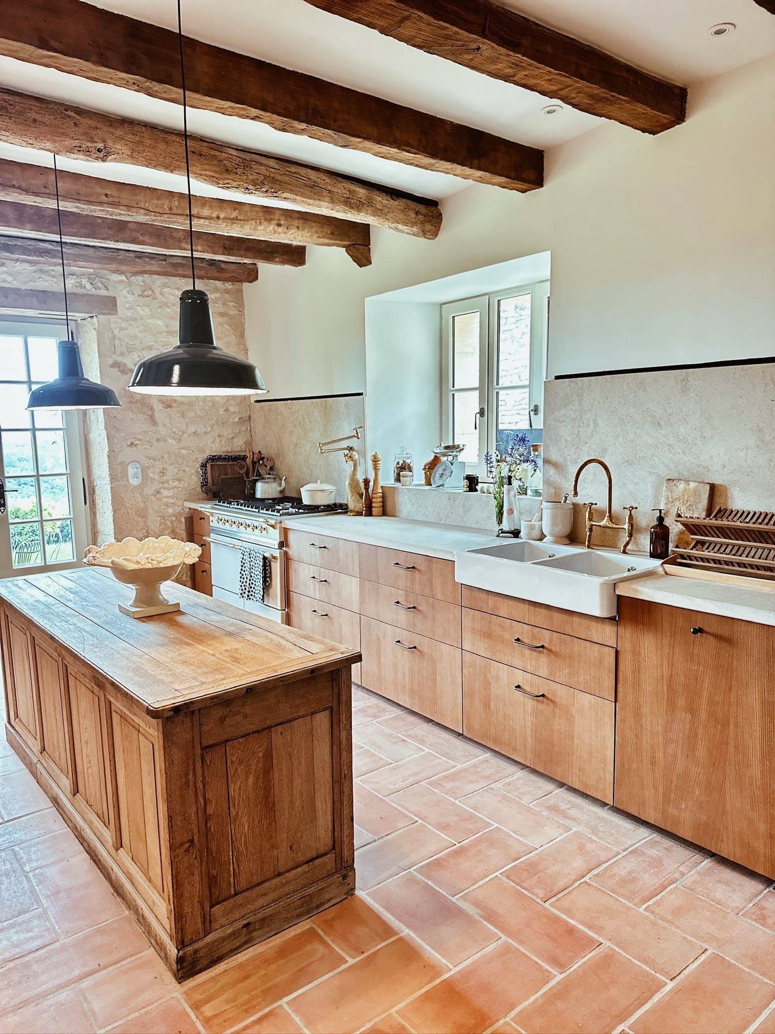 A rustic kitchen with wooden furniture, a farmhouse sink, exposed ceiling beams, a terracotta floor and a central wooden island. Sunlight streams in through a window and glass door, illuminating this warm space.