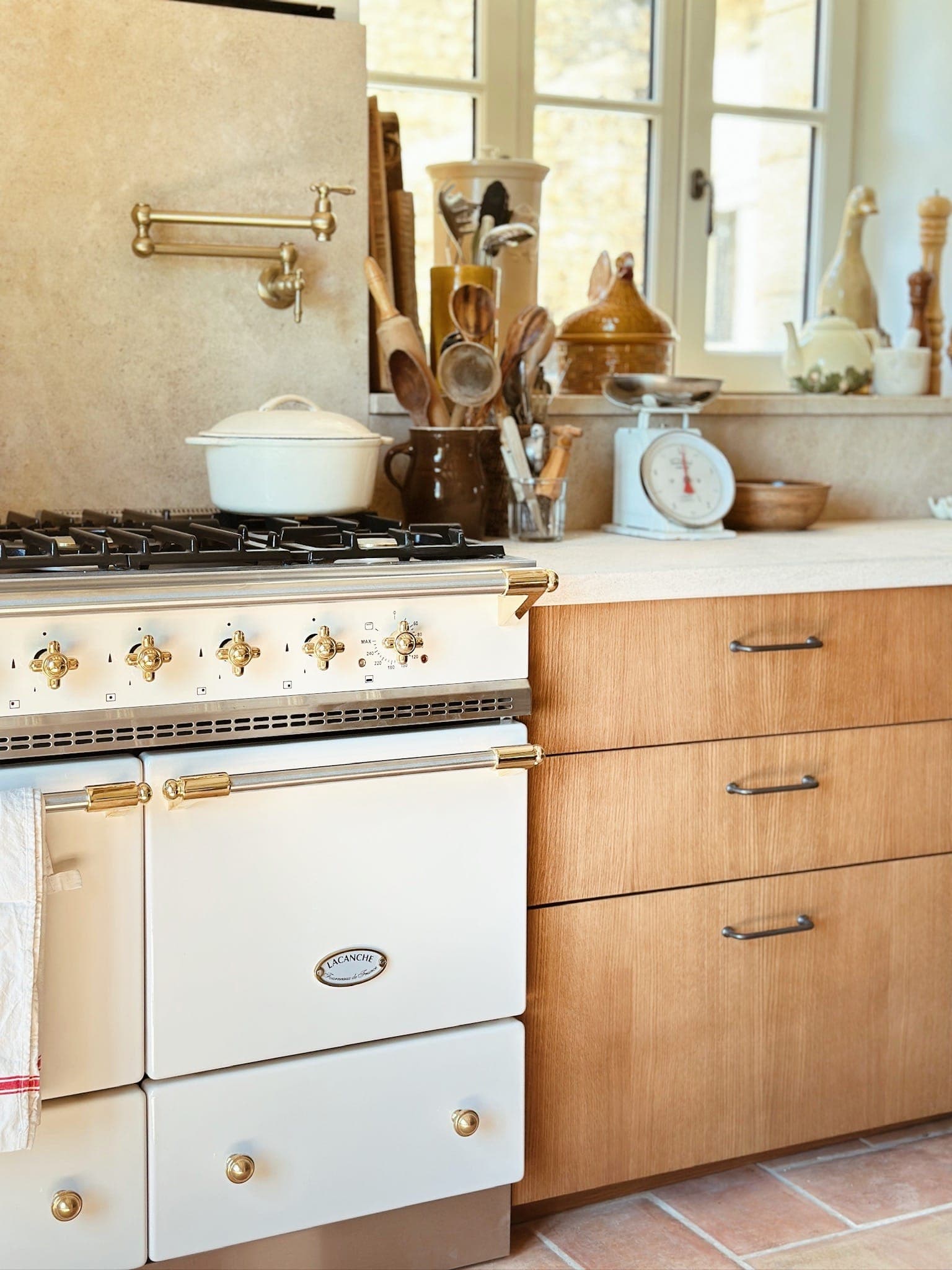 A vintage-style white stove with gold accents sits next to wooden cabinets in a warm kitchen. Utensils, jars and a white kitchen scale sit on the worktop beneath a window.