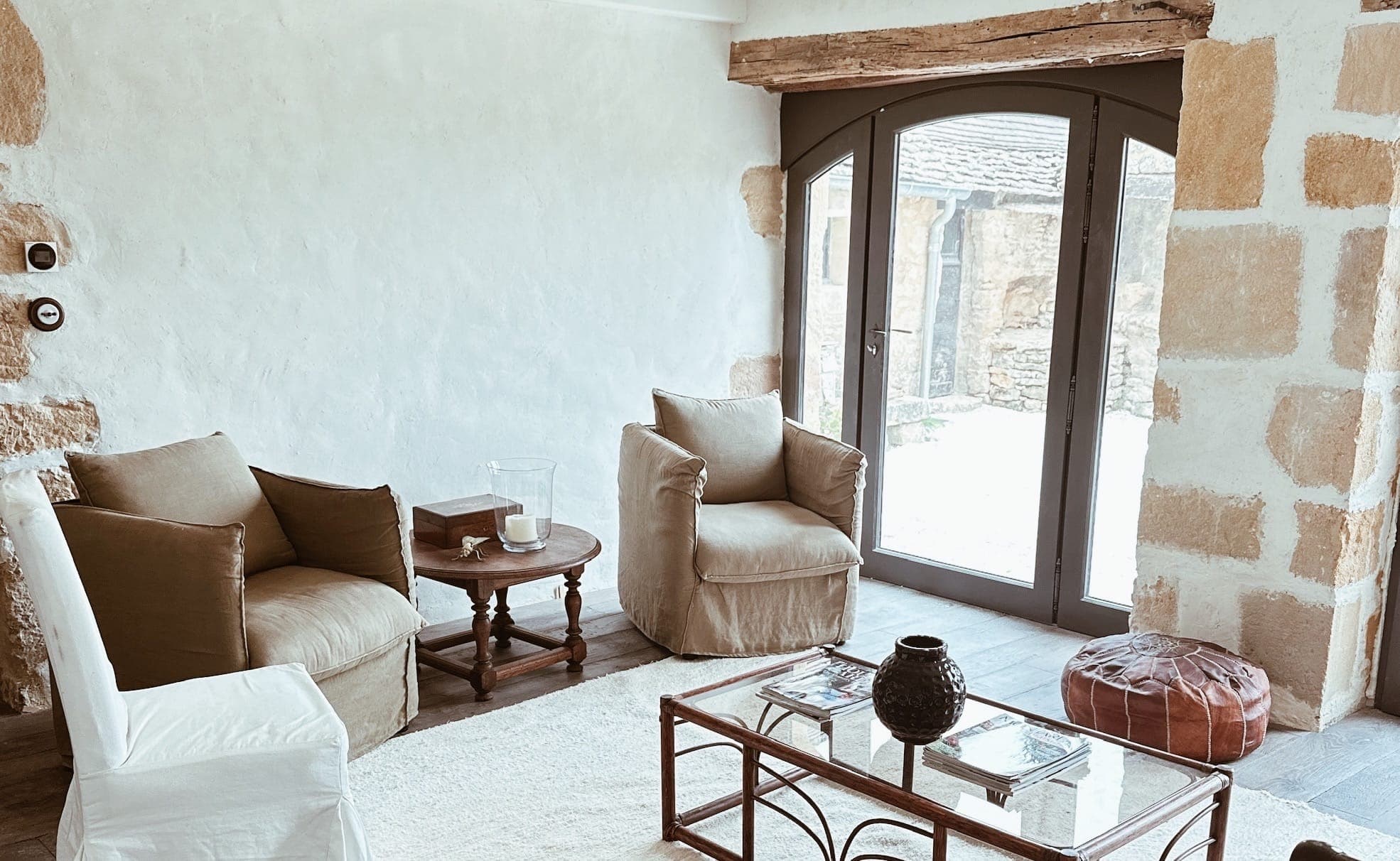 A warm living room with two beige armchairs, a small round wooden table, a glass coffee table, a brown pouffe and large curved glass doors letting in natural light. The walls are white, with exposed stonework and wooden beams.