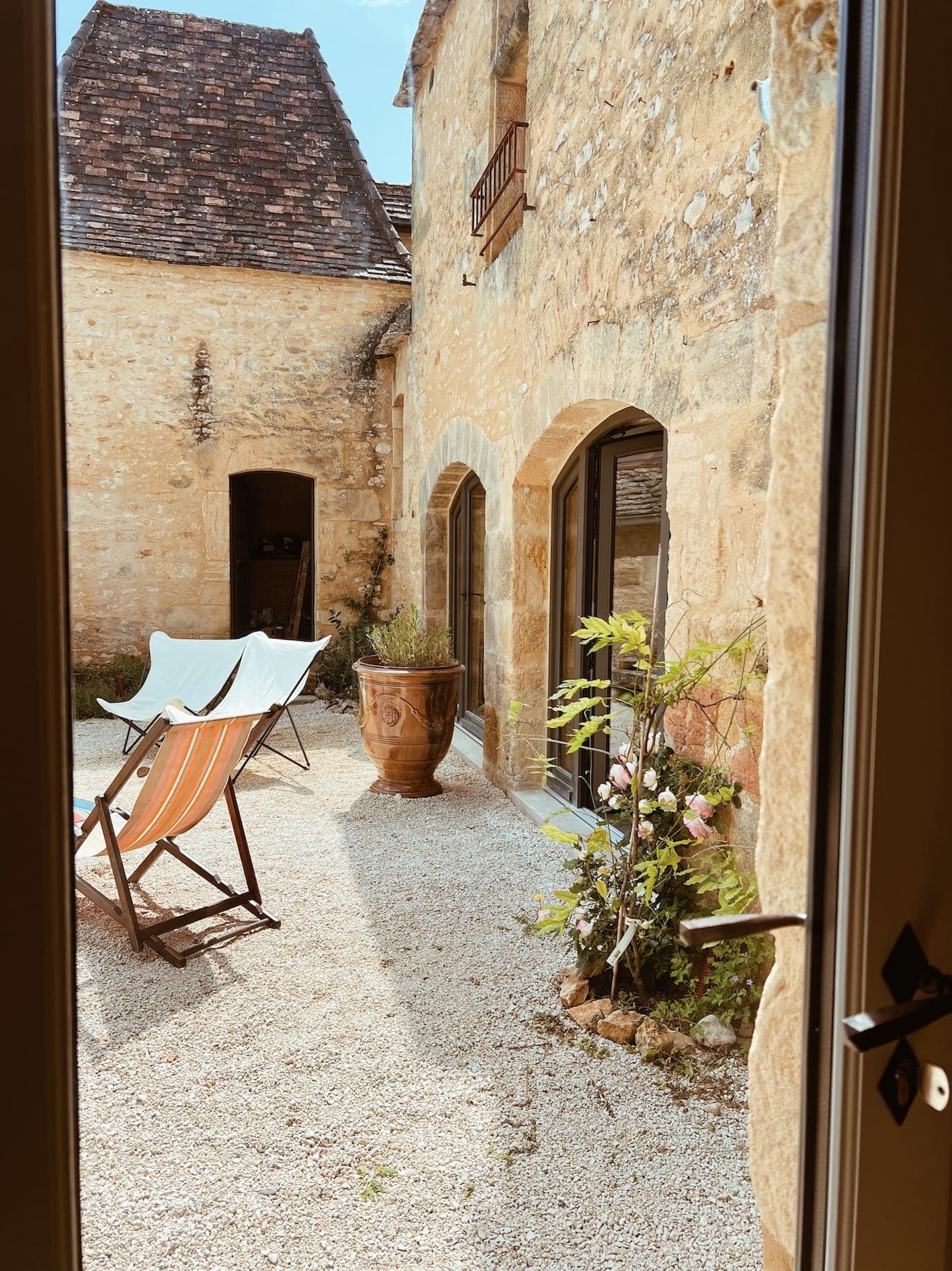 A sunny stone courtyard with two deckchairs, a potted plant, a gravel floor and rustic stone walls with arched doors and shuttered windows, seen from an open doorway.