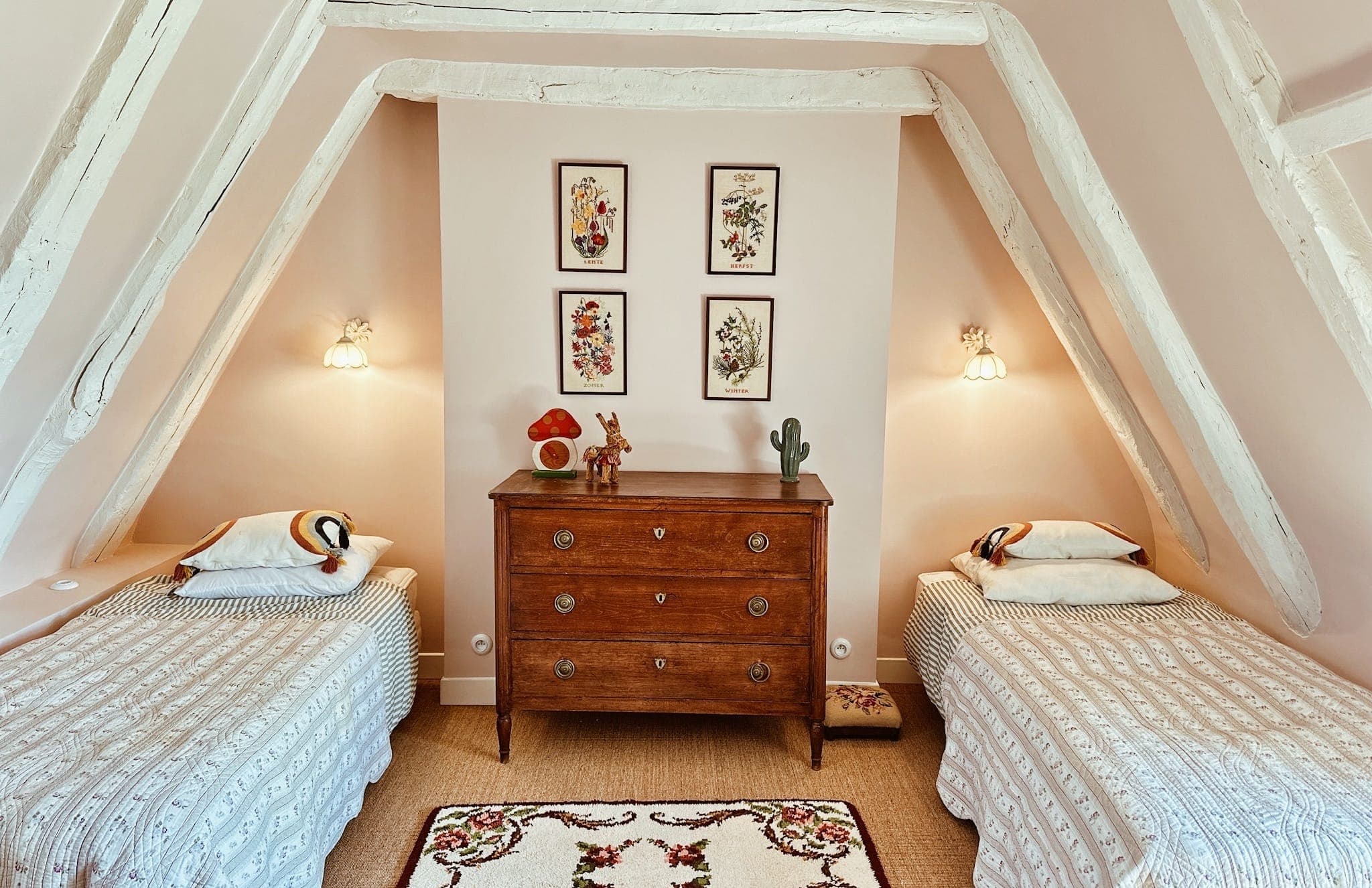 A cosy attic room with two single beds, a wooden chest of drawers in the middle, floral wall decorations, subdued lighting and exposed white beams. The beds have soft bedding and animal toys on the pillows.