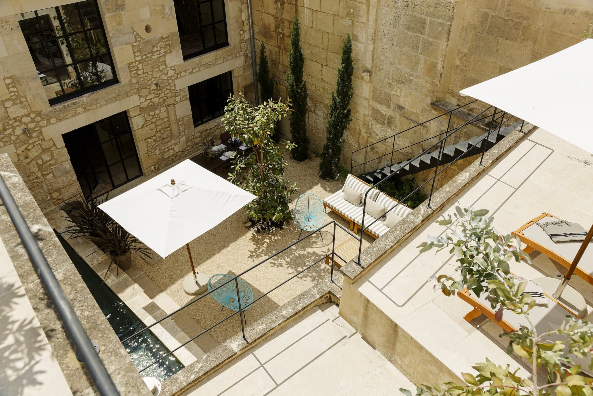 A sunny stone courtyard features a white parasol above a seating area with striped cushions, small round tables, green plants and deckchairs next to a narrow reflecting pool