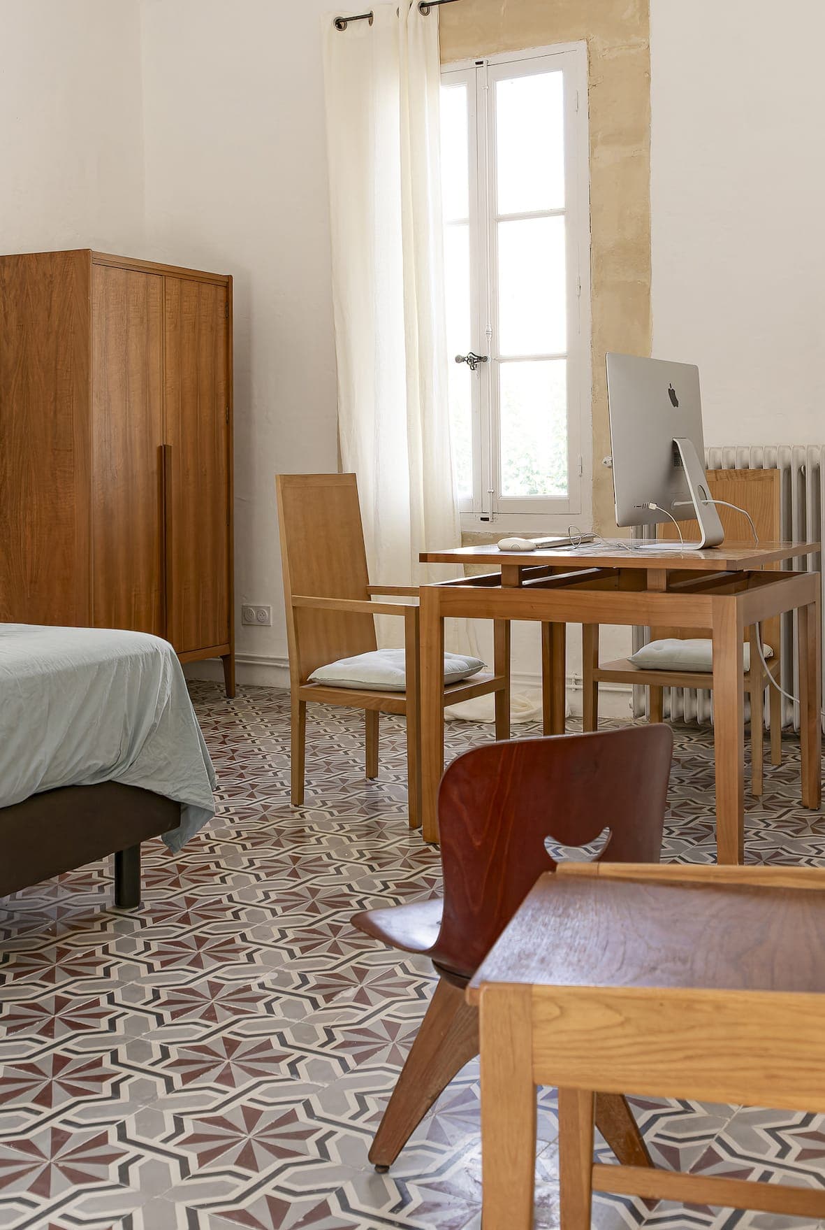 A bright room with a patterned tiled floor, wooden furniture, a bed with light bedding, a desk with an iMac, two wooden chairs, a wardrobe and a window with white curtains letting in natural light.