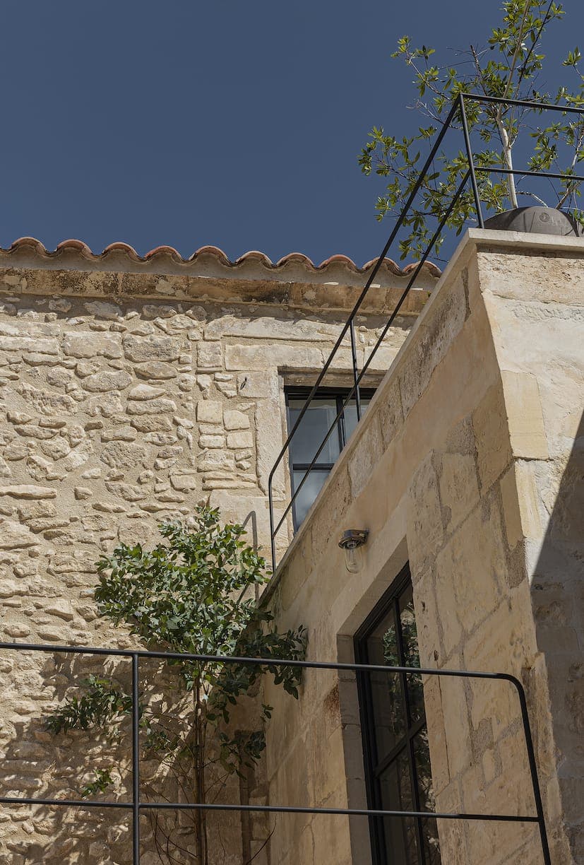 A stone building with a tiled roof and metal balustrades, with large windows and a small tree growing beside it under a clear blue sky.