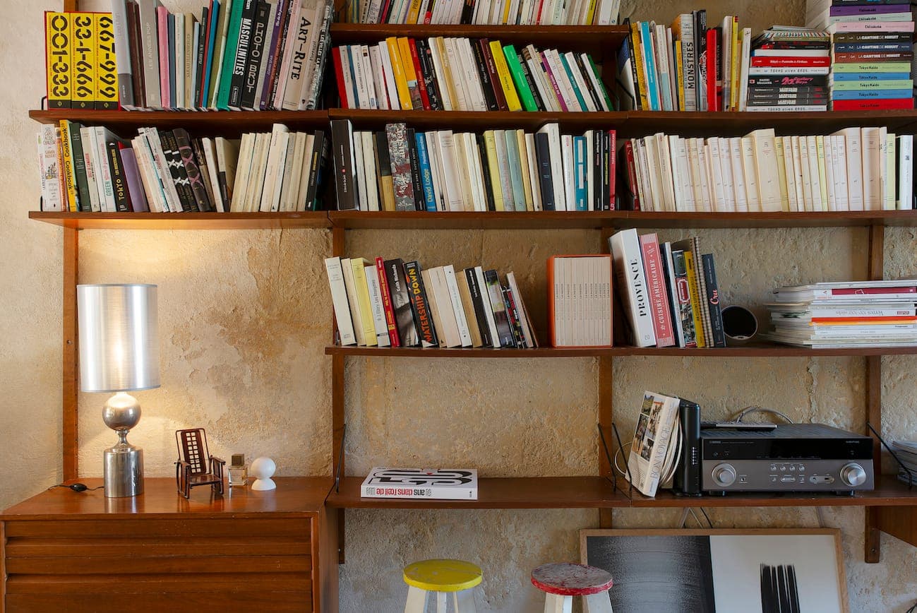 A cozy home office with wooden shelves filled with colorful books, a desk lamp, a small wooden desk, a sound system and two stools against a textured beige wall.