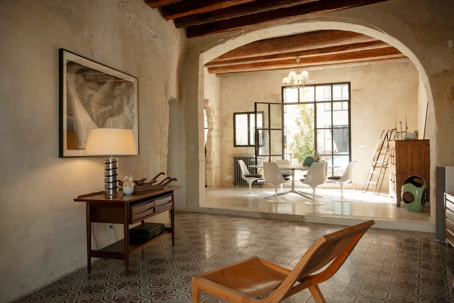 Salon et salle à manger lumineux avec murs en plâtre rustique, poutres apparentes, carrelage à motifs, mobilier en bois moderne, grande fenêtre et décoration minimaliste. Une lampe et des œuvres d'art sont posées sur une table d'appoint au premier plan.