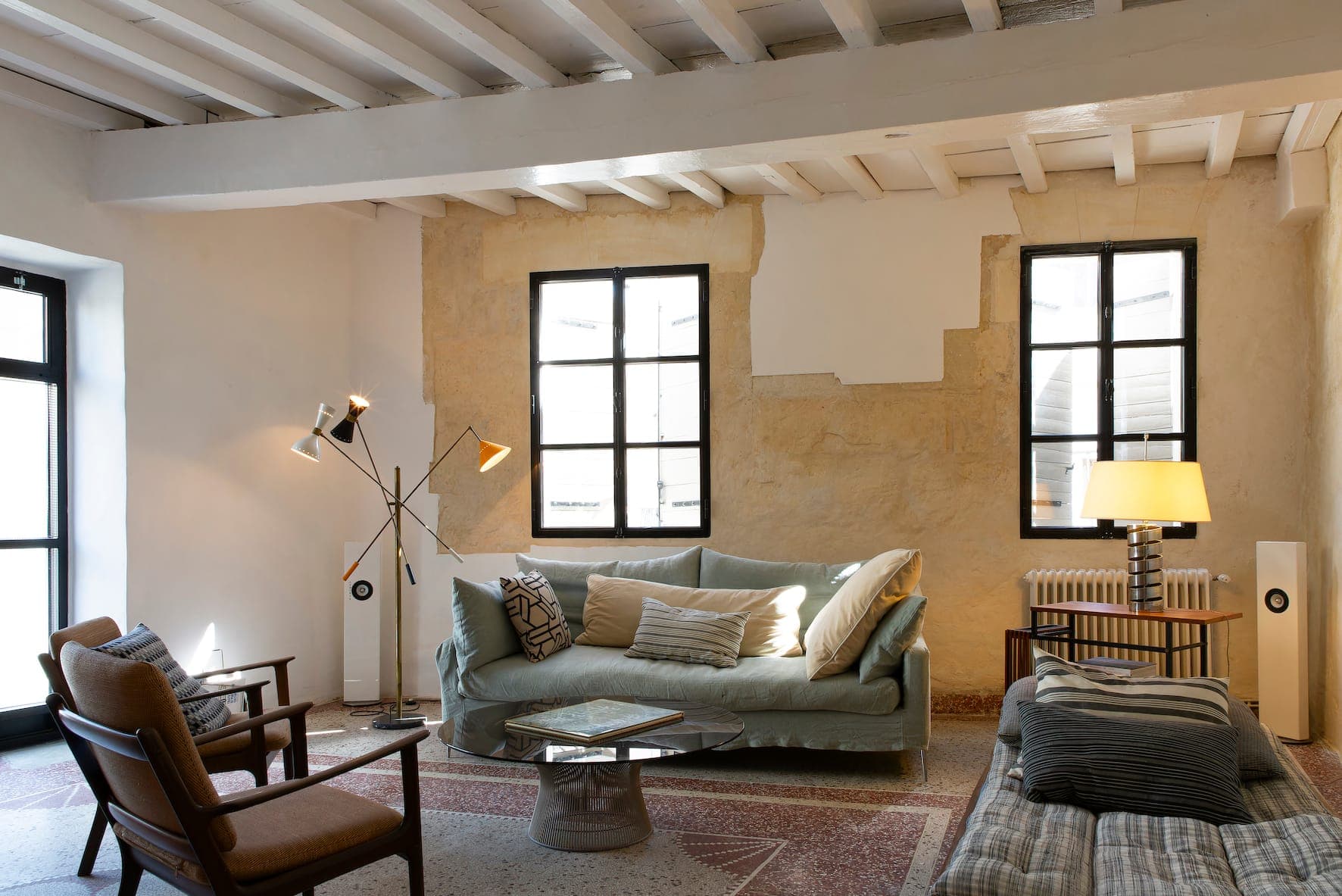 Bright, minimalist living room with white walls, exposed beams, comfortable sofa, wooden chair, small desk and natural light from large windows.