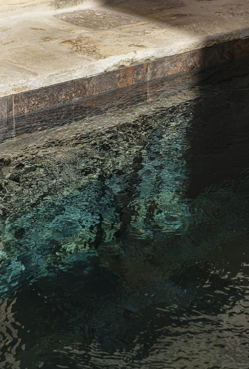 Sunlight reflects off the undulating surface of a clear-water pool, casting shadows and rippling patterns on a stone rim and submerged area.