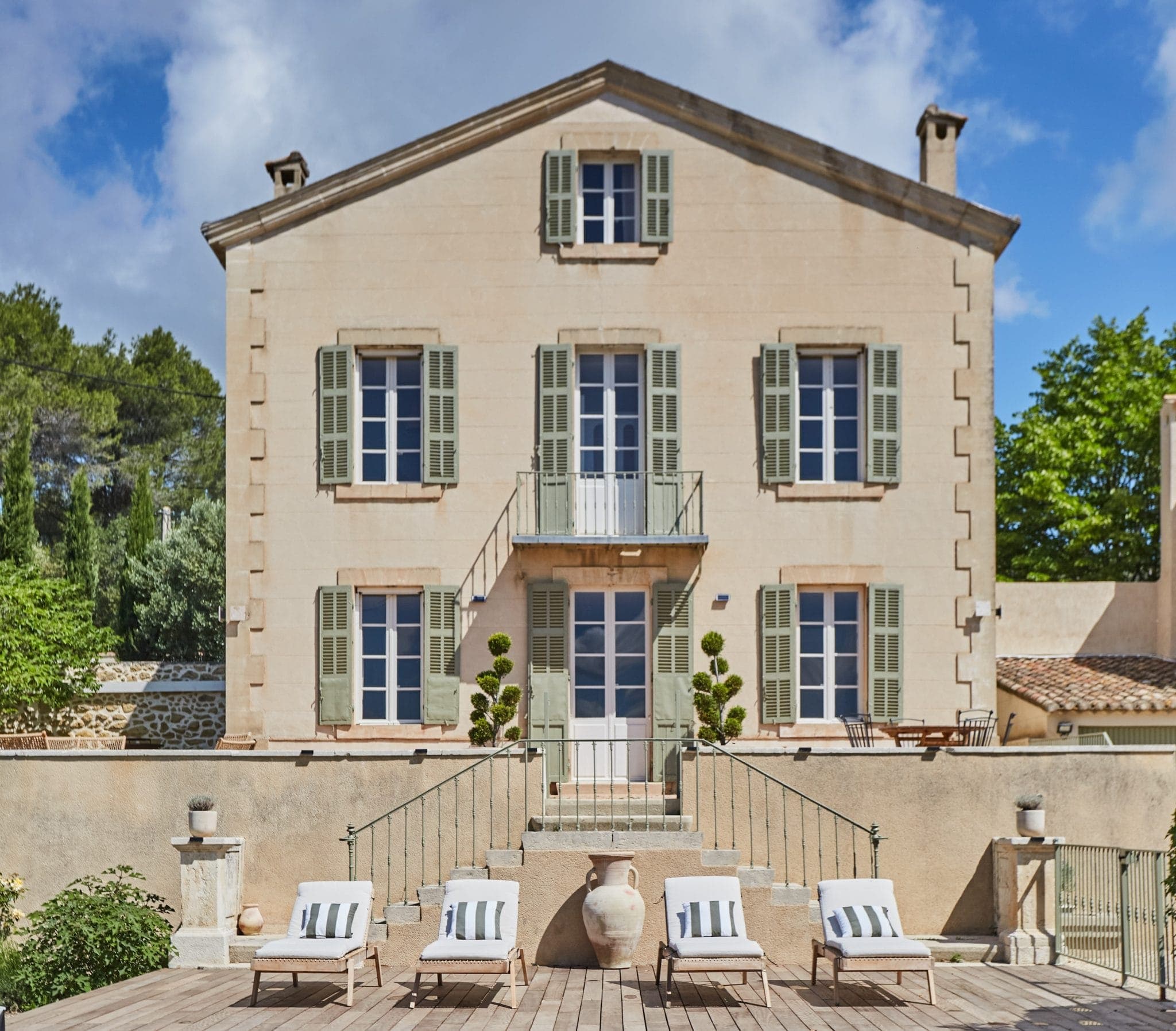 Une maison beige à deux étages avec des volets verts et un petit balcon, entourée de verdure. Devant, une terrasse avec quatre chaises longues aux coussins blancs et un grand vase décoratif.