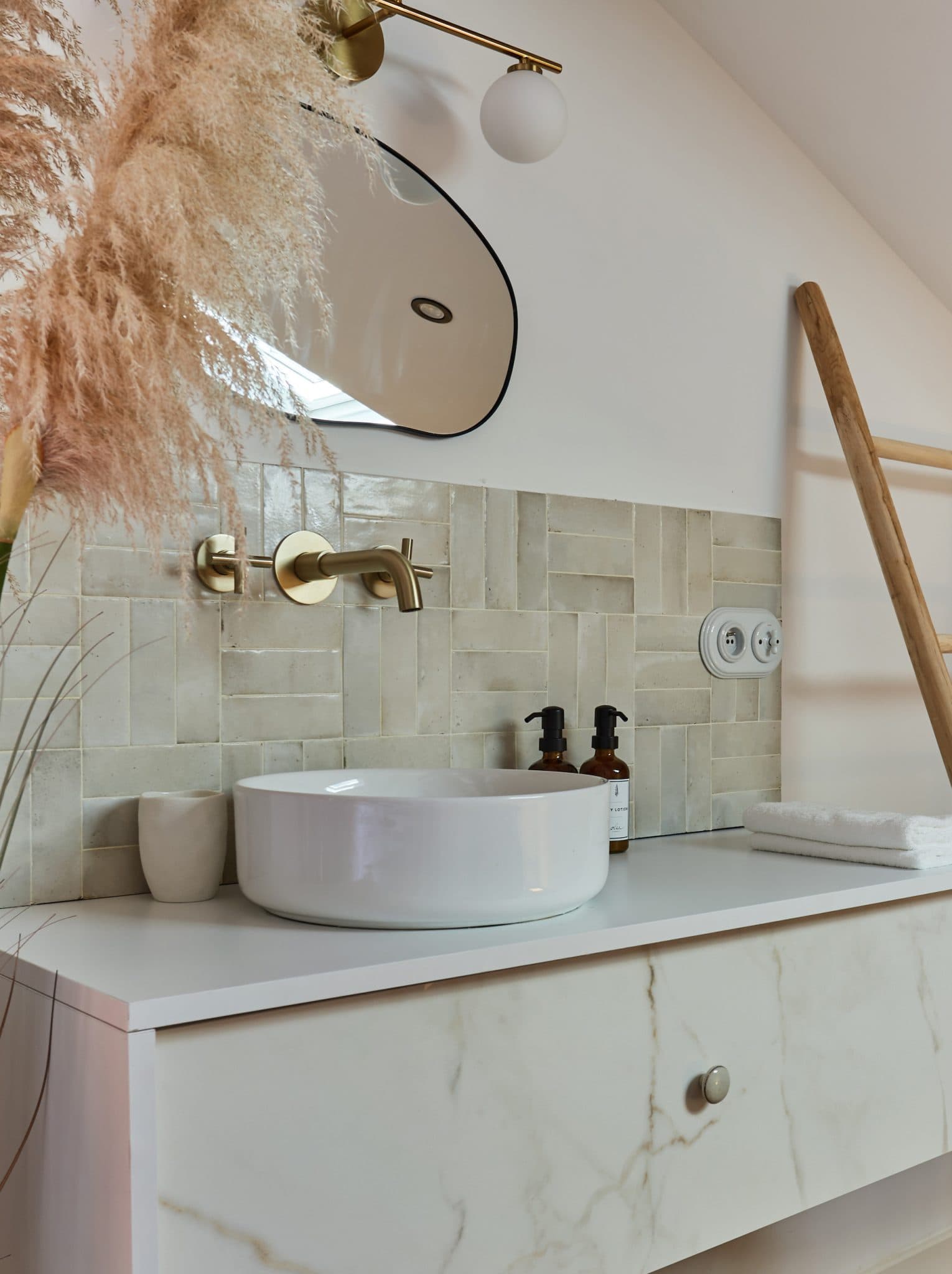 Salle de bain moderne, aux tons clairs et lumineux. Des produits de soin sont disposés à côté de l'évier et un grand miroir est accroché au mur.