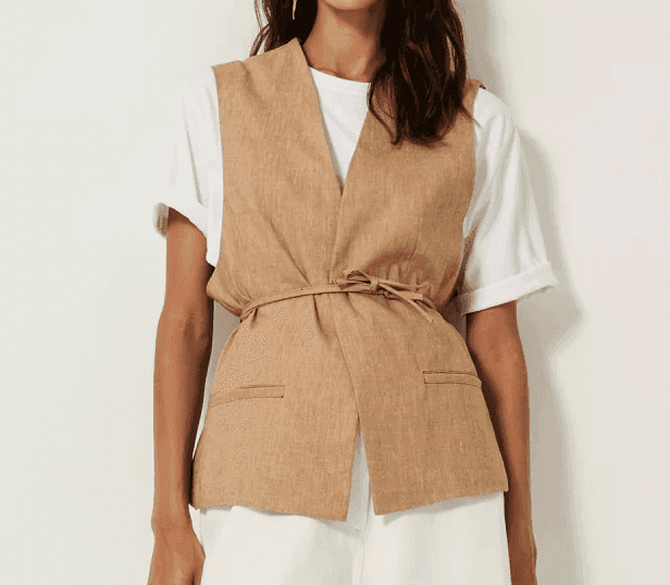 Femme portant un gilet beige sur un t-shirt blanc et un pantalon blanc. Elle se tient debout sur un fond uni et clair. Ses cheveux sont ondulés et elle porte une boucle d'oreille pendante.