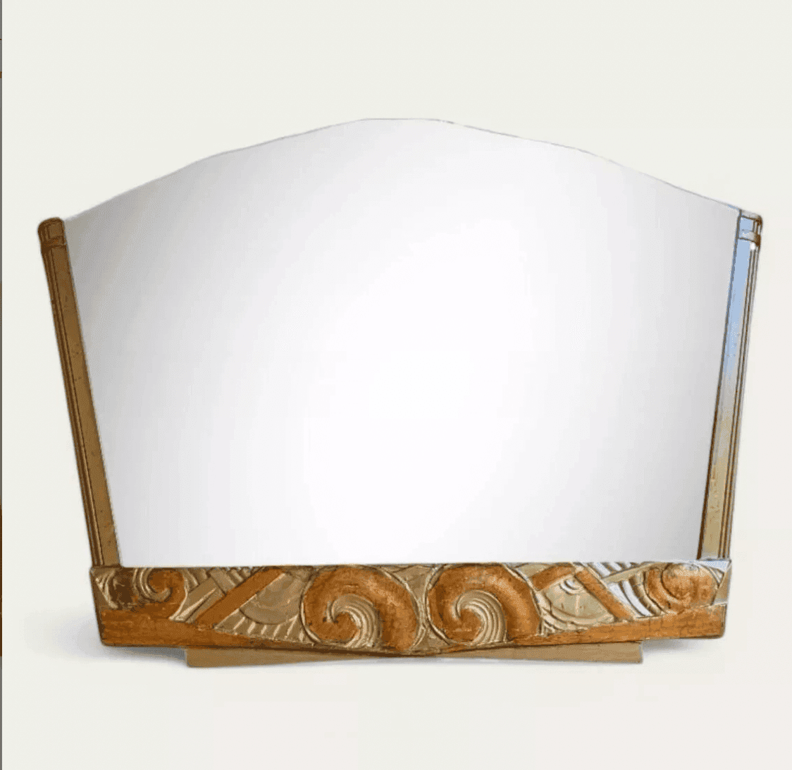 Un grand miroir vintage avec un dessus incliné et un cadre en bois présentant des motifs Art déco sculptés et ornés le long du bord inférieur.