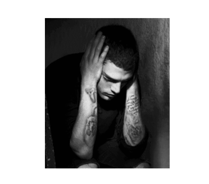 Un homme tatoué sur les bras est assis, les coudes sur les genoux, la tête entre les mains. Il semble angoissé ou plongé dans ses pensées, dans un décor monochrome et faiblement éclairé.