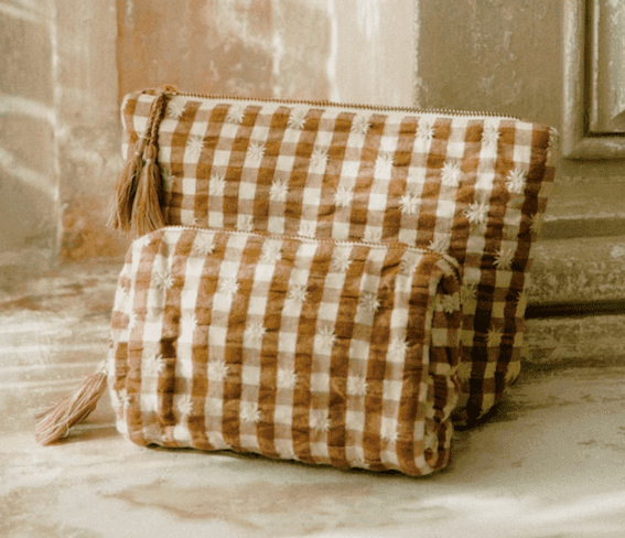 Deux pochettes zippées à pompons, en tissu doux et texturé à motif vichy marron et crème, sont placées sur une surface de couleur claire avec un fond patiné.