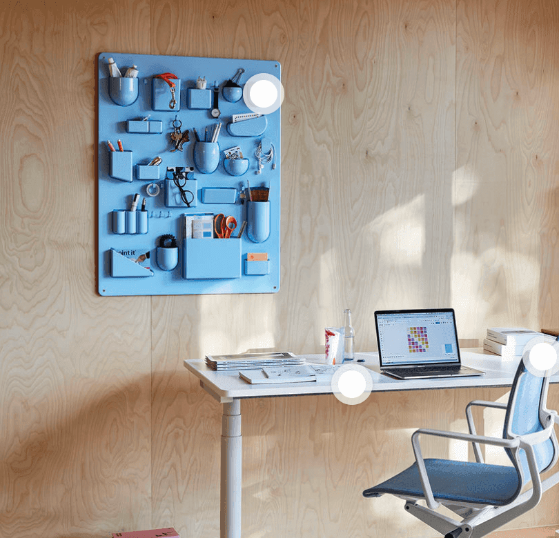 Un espace de travail bien rangé avec un bureau blanc, une chaise bleue rembourrée et un ordinateur portable. Au mur, un organiseur bleu clair contenant diverses fournitures de bureau se détache sur un fond en bois clair. La lumière du soleil projette des ombres sur la scène.