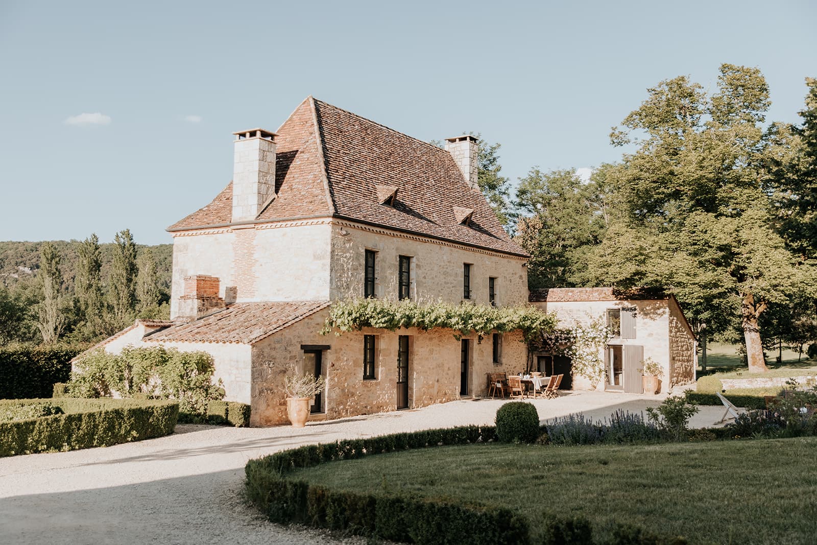 A charming stone house with a steep tiled roof, surrounded by manicured gardens and trees, sits under a clear blue sky. A small patio with outdoor furniture is visible near the entrance.