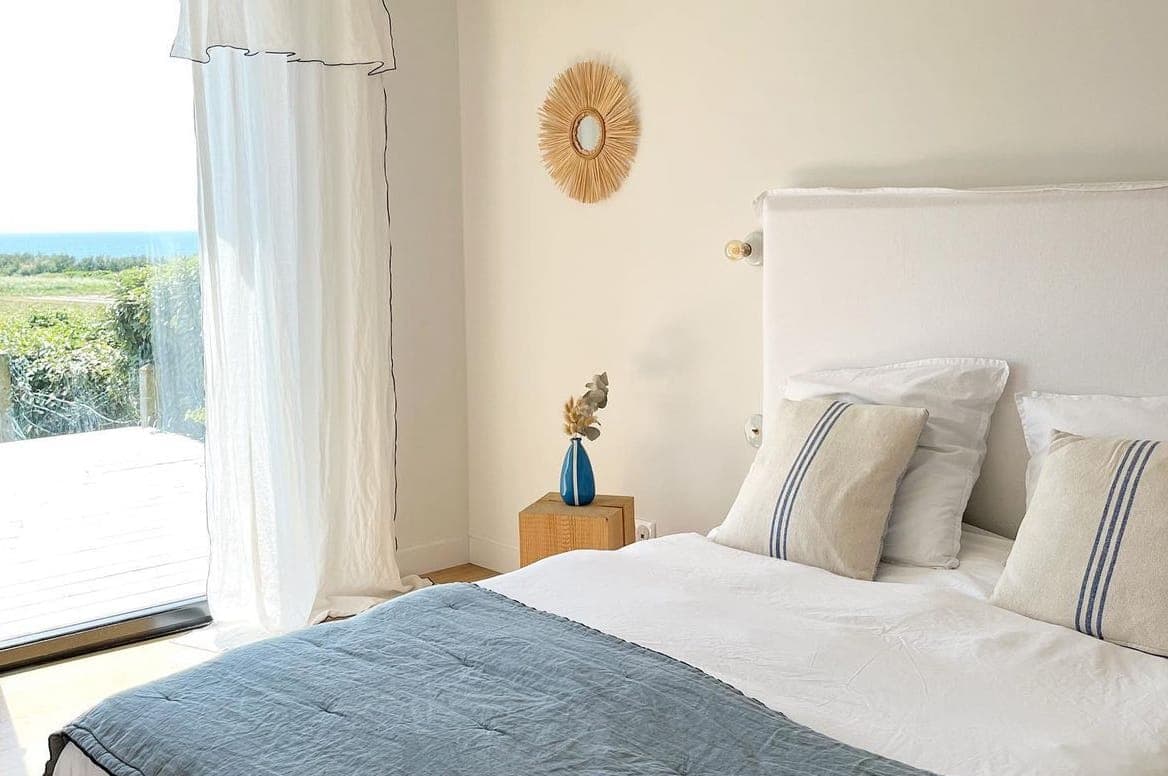 Une chambre lumineuse avec un grand lit avec des oreillers blancs et beiges, un jeté bleu clair, une table d'appoint en osier avec un vase bleu et des rideaux transparents laissant entrer la lumière du soleil par des portes vitrées donnant sur la verdure.