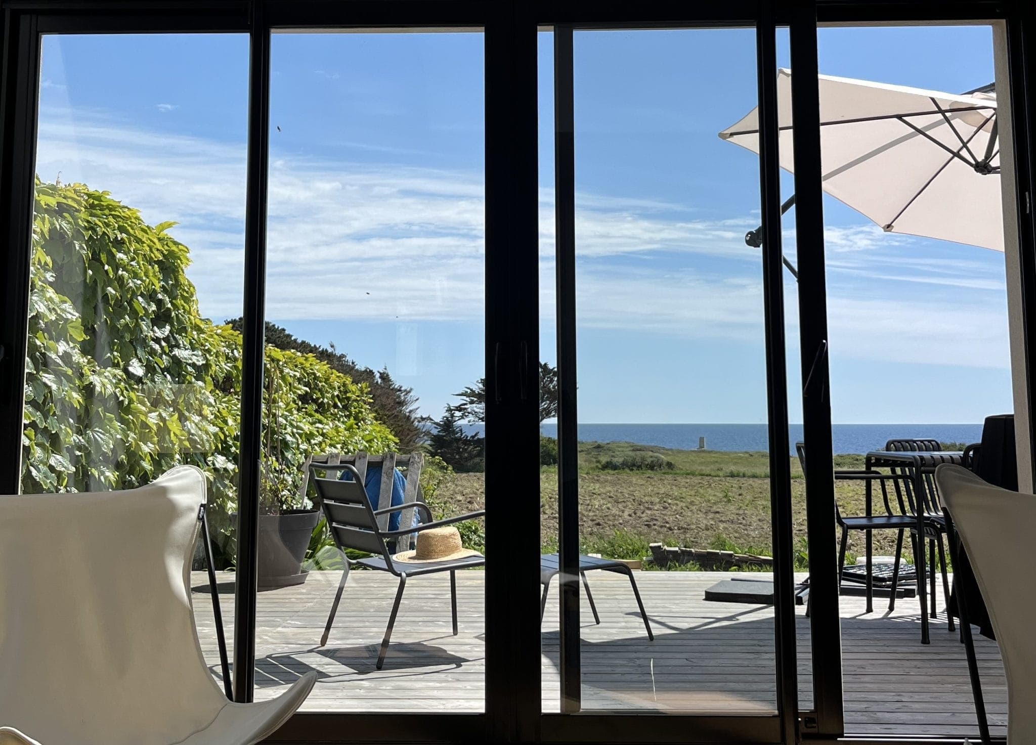 Vue à travers des portes vitrées sur un patio ensoleillé avec des chaises, un chapeau sur une chaise, des tabourets de bar, un grand parasol blanc, des plantes vertes et un horizon lointain de mer et de ciel bleus.