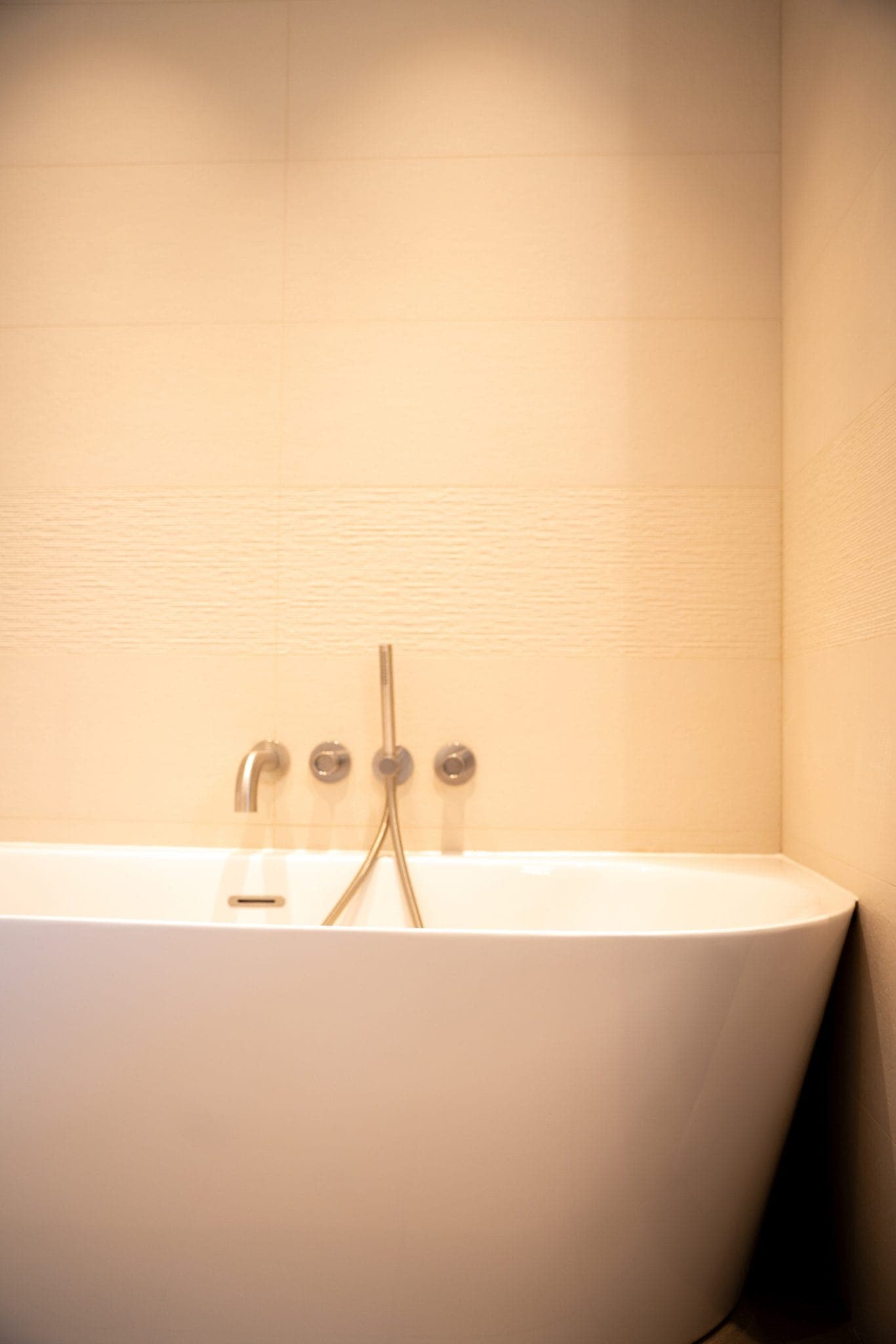 Baignoire blanche moderne avec des accessoires argentés élégants contre un mur carrelé beige dans une salle de bain doucement éclairée.