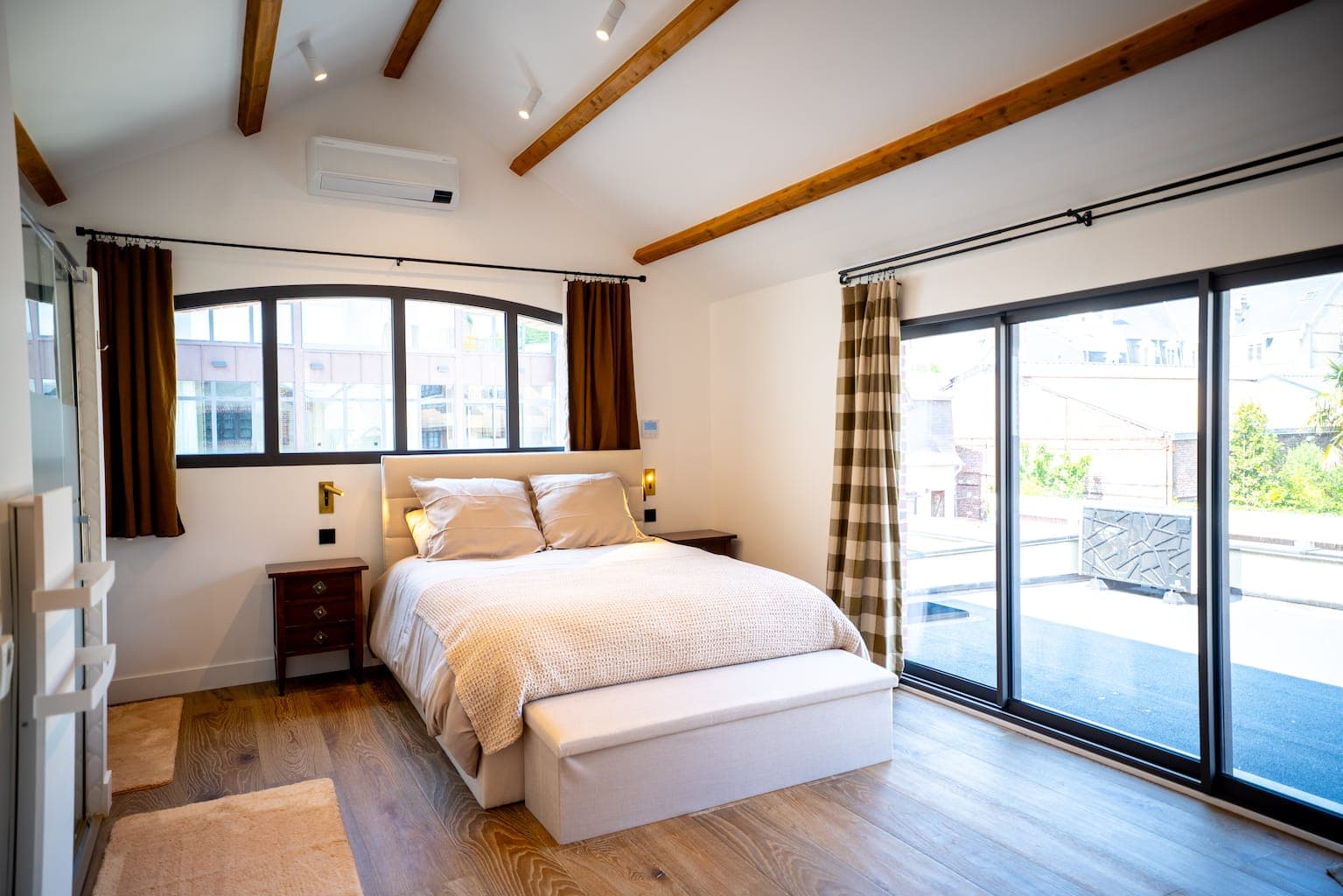 Une chambre lumineuse et moderne avec poutres apparentes, un grand lit, deux tables de chevet, du parquet et de grandes fenêtres avec rideaux laissant entrer la lumière naturelle. Une porte-fenêtre coulissante s'ouvre sur un balcon ou une terrasse.
