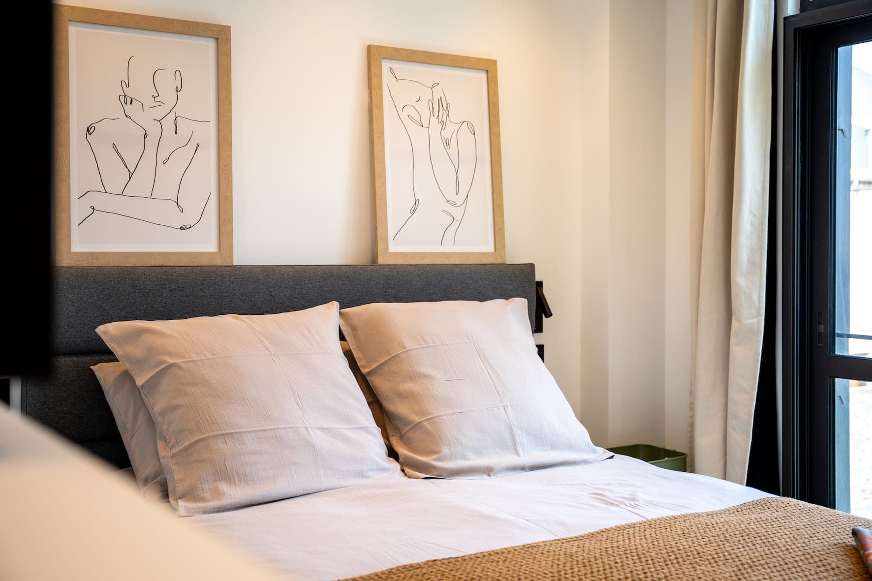 Un lit soigneusement fait avec des oreillers et une literie blancs se trouve sous deux portraits d'art minimalistes sur le mur dans une chambre lumineuse et moderne avec la lumière naturelle provenant d'une fenêtre voisine.
