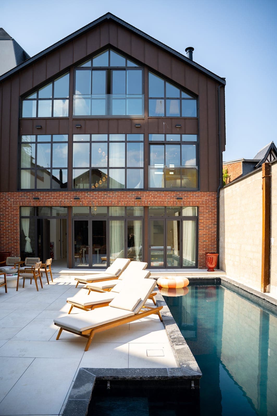 Une maison moderne de trois étages, dotée de grandes baies vitrées, donne sur une piscine. Plusieurs chaises longues en bois et une bouée orange ronde sont disposées sur la terrasse ensoleillée au bord de la piscine.