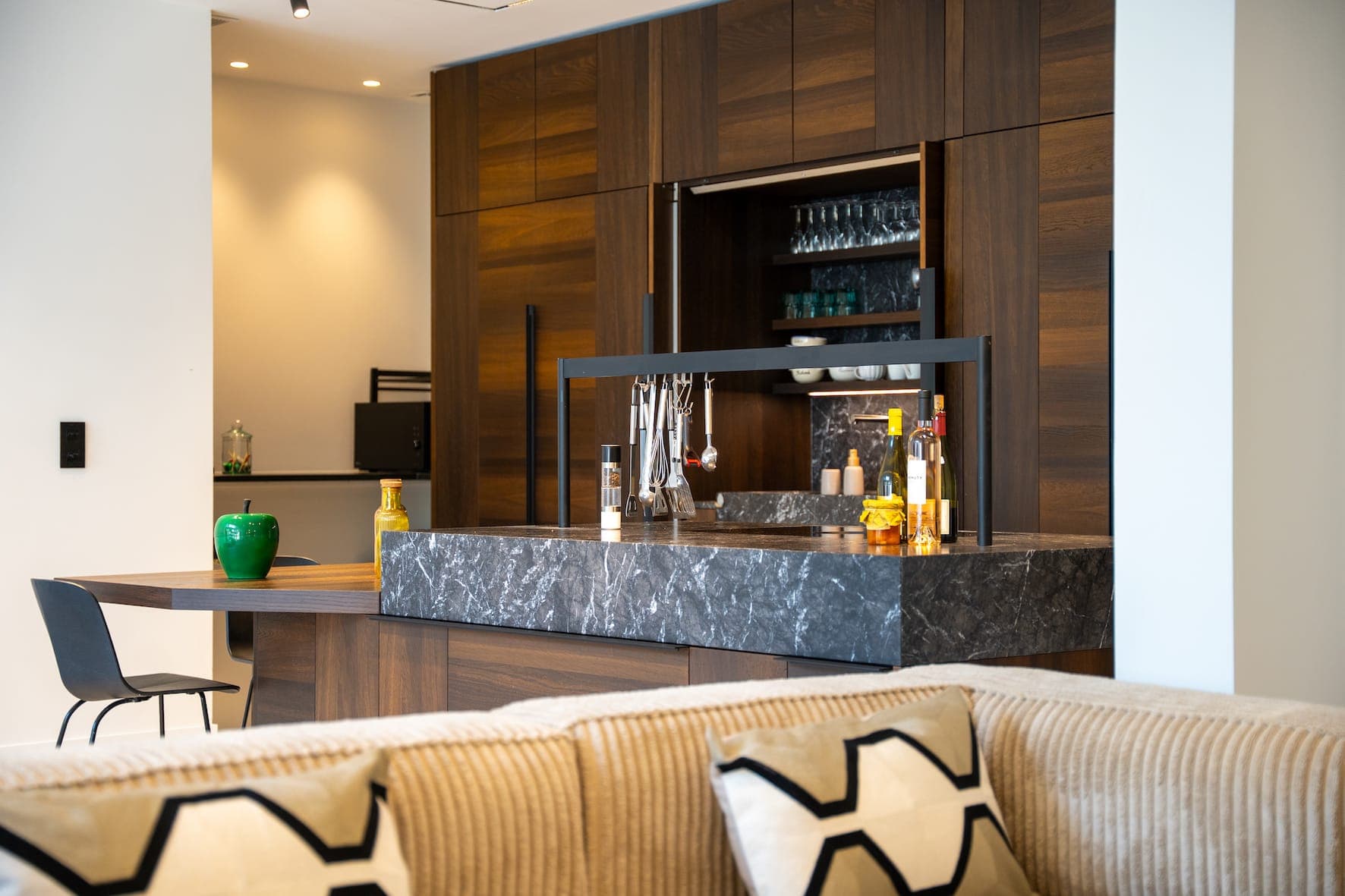 Cuisine moderne avec placards en bois foncé, îlot central en marbre noir, étagères ouvertes avec verres, bouteilles et objets de décoration. Au premier plan, un canapé beige avec des coussins à motifs géométriques.