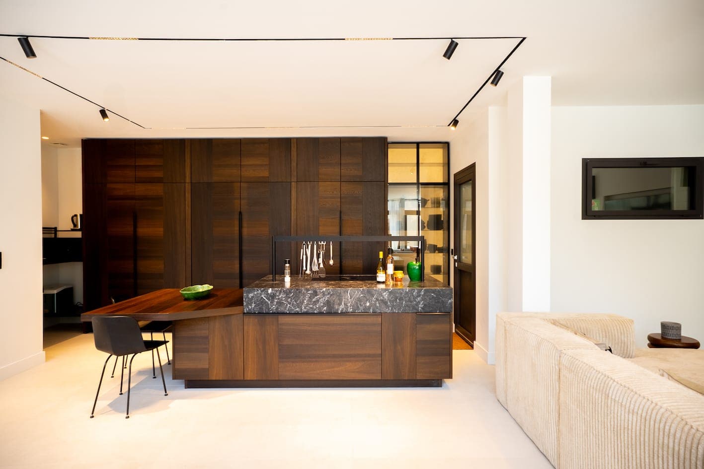 Cuisine moderne avec placards en bois foncé, îlot central en marbre, tabourets de bar et décoration minimaliste. À côté de la cuisine se trouvent un canapé beige et une télévision murale. Lumineuse et bien éclairée grâce à un éclairage sur rail.