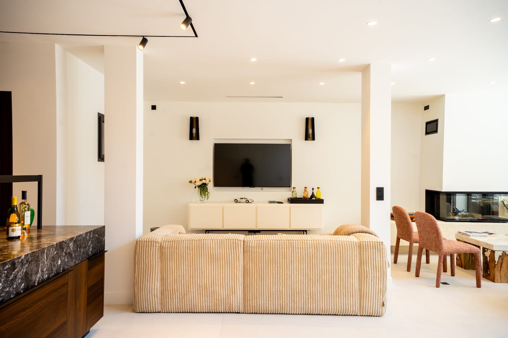 Un salon moderne avec un canapé en velours côtelé beige face à une télévision murale, une console multimédia blanche et des objets de décoration. À droite se trouve un coin repas avec une table en verre et des chaises roses. L'espace est lumineux et affiche une décoration minimaliste.