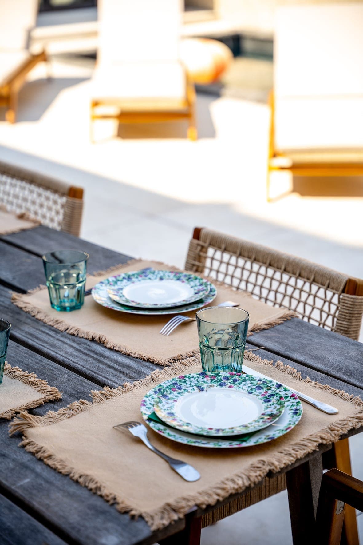 Une table à manger d'extérieur est dressée avec des assiettes, des bols et des verres fleuris posés sur des sets de table beiges. Des chaises à dossier tressé entourent la table, et des fauteuils de salon flous se dessinent en arrière-plan ensoleillé.