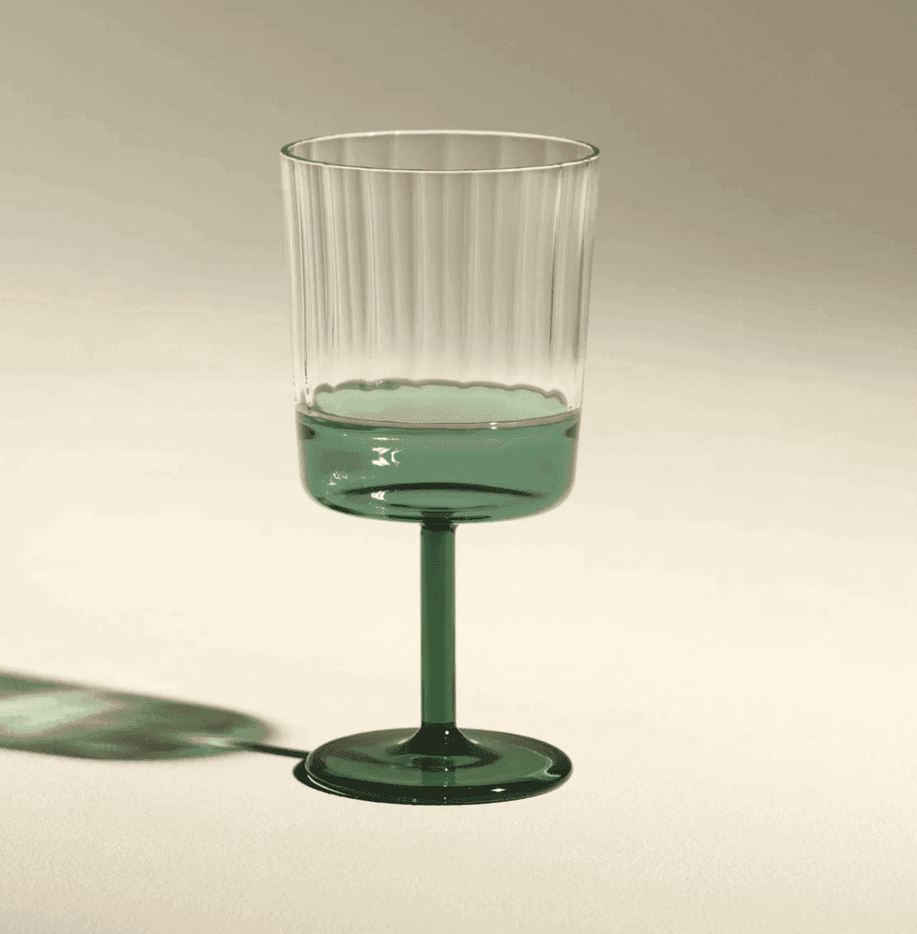 Un gobelet en verre transparent avec des crêtes verticales, comportant une base, une tige et un bol inférieur teintés de vert, posé sur une surface beige clair avec une ombre douce à gauche.