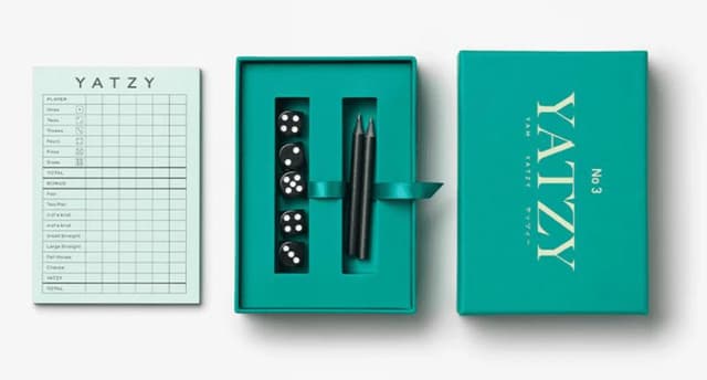Coffret bleu sarcelle avec "Yatzy No. 3" sur le couvercle. A l'intérieur : cinq dés noirs à pois blancs et deux stylos. Un bloc-notes avec les détails du jeu est placé à côté de la boîte.