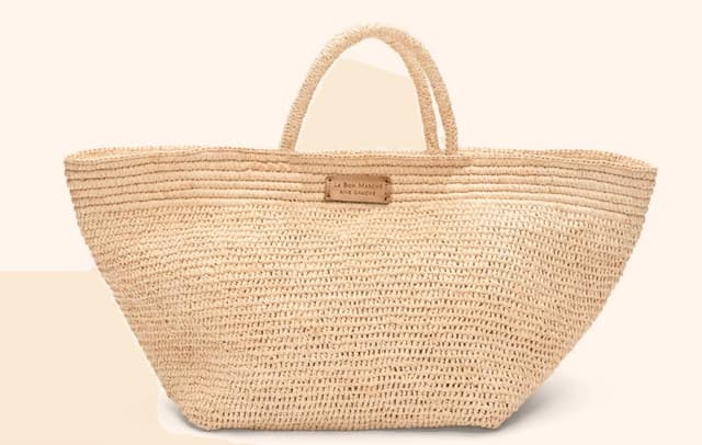 Un grand sac fourre-tout en paille tressée avec deux anses et un petit empiècement en cuir affichant un texte sur le devant. Le sac se présente sur un fond beige uni.