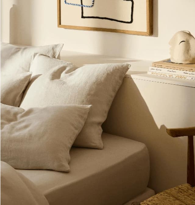 Un coin chambre douillet avec un lit aux oreillers et draps beiges et blancs moelleux. Une petite table de nuit en bois contient des livres empilés et un objet de décoration fantaisiste en forme de chat. Au-dessus, une œuvre d'art abstraite encadrée est accrochée au mur.