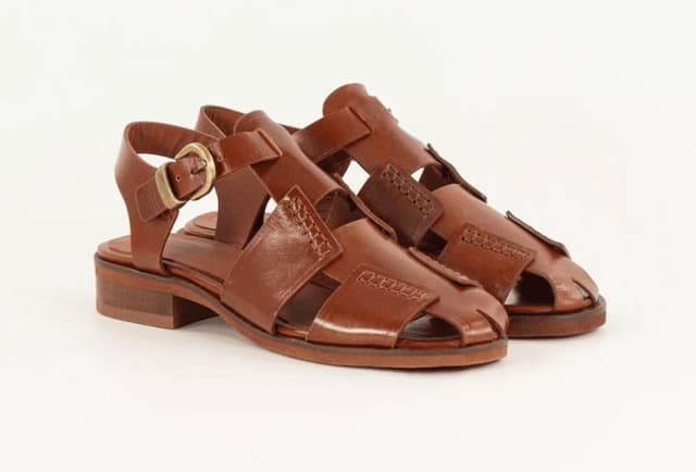 Une paire de sandales en cuir marron à bout fermé, dotées d'une bride à boucle, de détails cousus et d'un talon bas bottier. Elles sont positionnées sur un fond blanc uni.