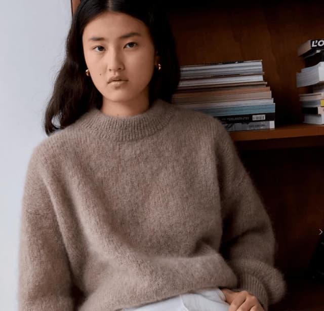 Une personne aux cheveux longs et foncés porte un pull marron confortable et surdimensionné et un pantalon blanc, assise devant une étagère en bois remplie de livres empilés. Elle a une expression neutre et regarde directement la caméra.