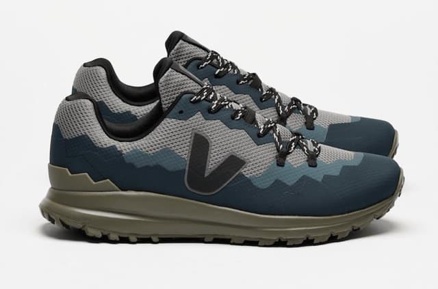 Une paire de baskets élégantes au design camouflage dans des tons bleus et gris. Elles sont dotées de lacets complexes et d'une semelle robuste vert olive, sur fond blanc uni.