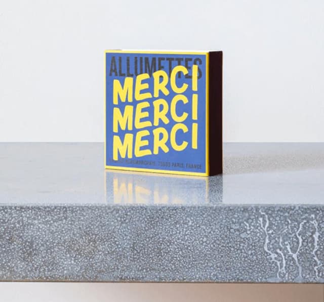 Une boîte carrée d'allumettes portant l'inscription « ALLUMETTES MERCI MERCI MERCI » en lettres jaunes audacieuses sur fond bleu repose sur une surface grise mouchetée. Au fond de la boîte, on peut lire « 111 Boulevard Beaumarchais, 75003 Paris, France ».