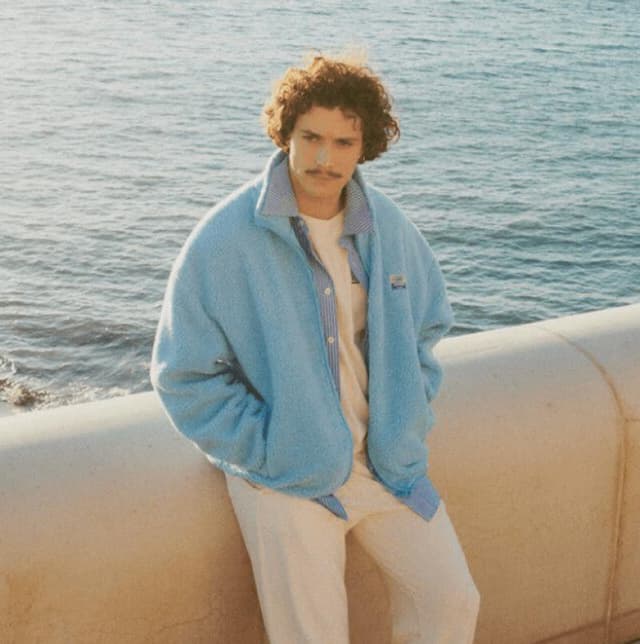 Un homme aux cheveux bouclés et à la moustache, vêtu d'une veste bleu clair et d'un pantalon blanc, s'appuie contre un mur de pierre au bord de la mer. L'océan est calme et le soleil se couche, projetant une lumière chaude sur la scène.