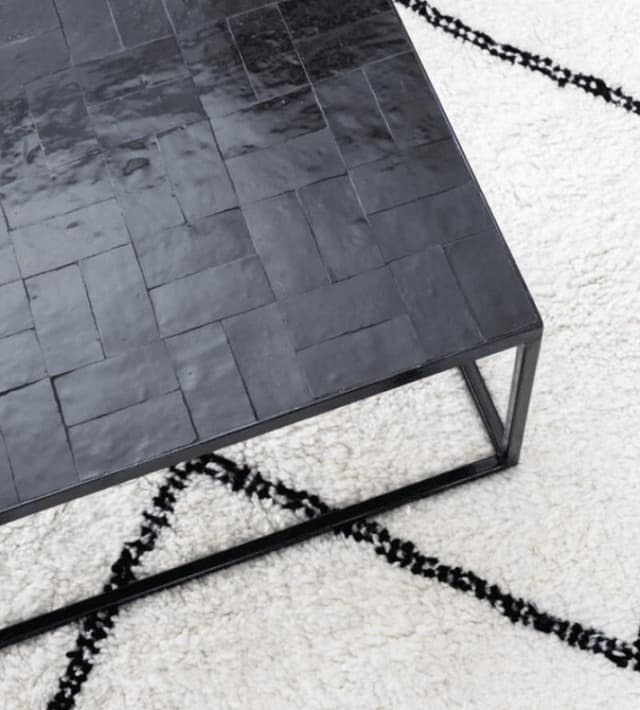 Une table basse noire brillante, ornée d'un motif de carrelage, est posée sur un tapis blanc aux motifs géométriques noirs. La table est dotée d'une structure métallique simple et sa surface reflète subtilement la lumière.