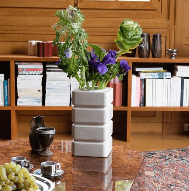 Un vase blanc moderne orné de feuillage vert et de fleurs violettes est posé sur une table en bois. À proximité se trouvent deux tasses à expresso, un petit pichet et une assiette de raisins. Une bibliothèque bien garnie se trouve à l'arrière-plan.