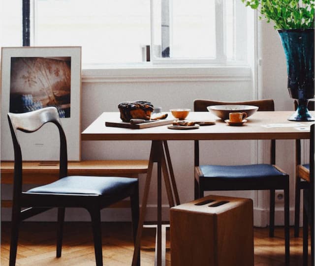 Un coin repas chaleureux avec une table en bois dressée pour le thé. On y trouve des tasses, une miche de pain et des fruits sur des plateaux. Un vase bleu garni de verdure, une œuvre d'art encadrée sur un banc et des chaises en bois sur un parquet. La lumière pénètre par une fenêtre.