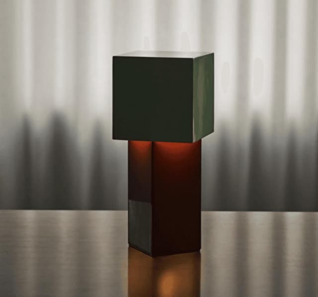 Une lampe carrée moderne et minimaliste, dotée d'un abat-jour vert foncé et d'une base sombre, repose sur une surface réfléchissante. Une douce lumière jaillit de l'intérieur, projetant des ombres subtiles sur un fond gris flou.